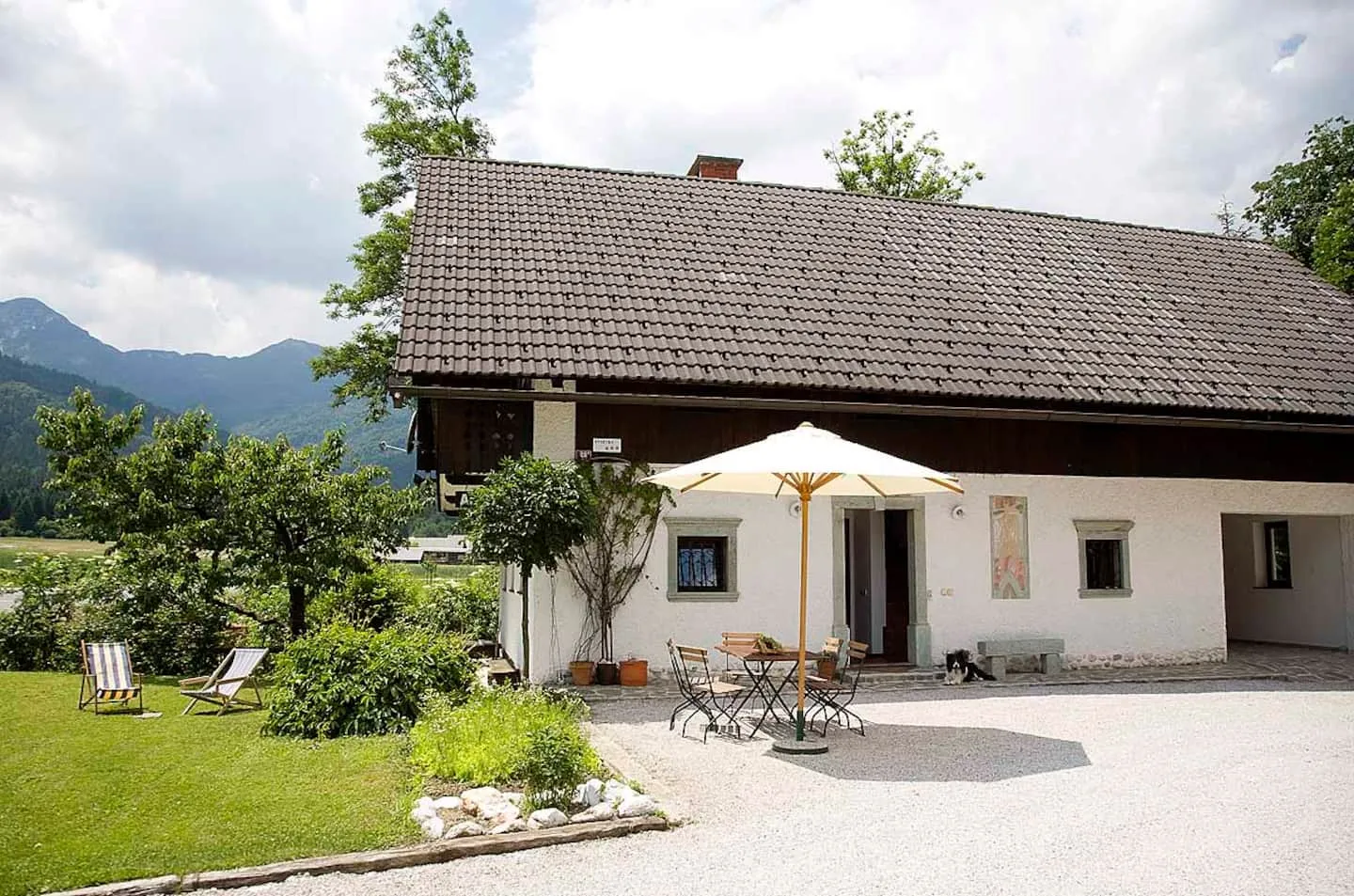 Bela hiša z apartmaji Pristavec s senčnikom na dvorišču, za njo gore
